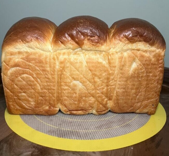 side view of a golden milk bread sandwich bread with three sections and darker brown top compared to the bottom resting on a yellow silicone mat on top of a wooden surface